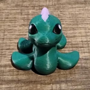 May include: A small, 3D-printed figurine of a green creature with a smooth, rounded body, large black eyes with white highlights, and a single light purple horn on its head. The surface shows horizontal layer lines from the printing process.
