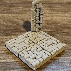 May include: A light gray miniature dungeon terrain piece. The base is a square of cobblestones, and a vertical pipe structure rises from the center. The piece is designed for tabletop gaming.