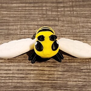 May include: A handcrafted bee figurine with a yellow body, black stripes, and large black eyes. The bee has white wings and black legs. The figurine is set against a wooden background.