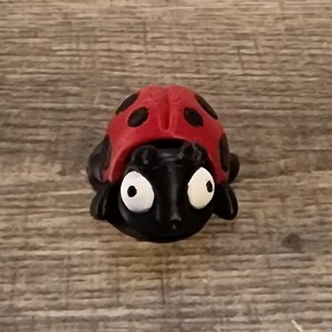 May include: A small, black ladybug figurine with a red shell and black spots. The ladybug has large, white eyes and is set against a wooden background. The figurine is a decorative item.