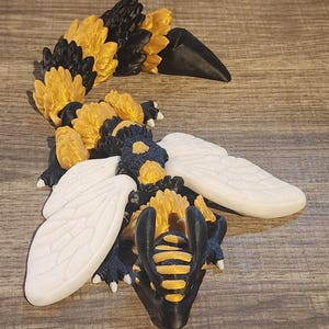 May include: A 3D printed articulated creature featuring a black and gold segmented body with a long, flexible tail. The figure has two large, smooth white wings extending from the sides and a black head with gold accents and small white claws.