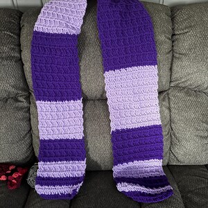 May include: A handmade crochet scarf in shades of purple. The scarf features alternating stripes of light and dark purple, creating a textured pattern. The scarf is displayed on a gray surface.