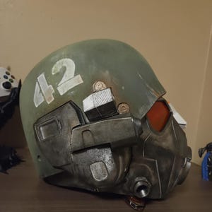 May include: A tactical helmet featuring a matte olive green dome with the number 42 painted in white on the side. The lower section is constructed from weathered metallic grey components, including a prominent orange visor and a circular port.
