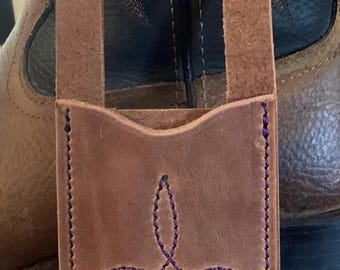 Op de afbeelding: Een bruin lederen tasje met twee verticale bandjes voorzien van metalen drukknopen aan de bovenkant. Het tasje heeft paarse stiksels langs de randen en een decoratief paars gestikt patroon op de voorzijde, inclusief een centraal motief en twee lijnen.