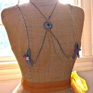 May include: A body chain with a dark metal chain, a central iridescent ring with a black square, and two iridescent clamps. The chain is displayed on a burlap mannequin with a brown ribbon.
