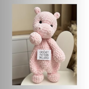 May include: A light pink crocheted hippo stuffed animal with small black eyes and small ears. The hippo is holding a small ball-shaped object. A tag reads "CROCHET PATTERN INCLUDED".