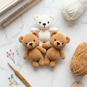 May include: Three crocheted teddy bears in brown and white, with black button eyes and stitched noses. A crochet hook and pins are next to the bears. A ball of white yarn and a ball of brown yarn are also in the image. Books are in the background.