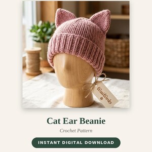 May include: A dusty pink knitted beanie with two pointed cat ears on top, featuring a ribbed cuff. The beanie is displayed on a wooden mannequin head with a small beige paper tag attached by a string that reads WasiWorks with a paw print.