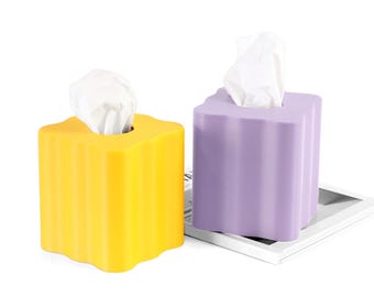 May include: Two decorative tissue box holders. One is yellow, the other is lavender. Both have a scalloped edge design and white tissues protruding from the top. The boxes are on a white background.