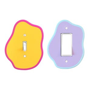 May include: Two decorative light switch covers. One is yellow with a pink border, the other is lavender with a light blue border. Both have a unique, organic shape and a silver toggle switch.