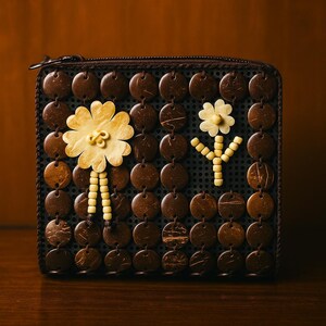 Könnte beinhalten: Eine braune Geldbörse mit floralem Design. Die Geldbörse besteht aus kleinen, runden, dunkelbraunen Teilen mit zwei Blumenmustern aus hellgelben und cremefarbenen Perlen. Die Geldbörse hat einen Reißverschluss.
