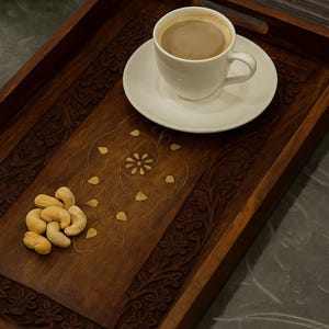 Könnte beinhalten: Ein dunkelbraunes Holzserviertablett mit geschnitzten Blumenmustern und einem Griff. Es enthält eine weiße Tasse und Untertasse mit einem kaffeeähnlichen Getränk sowie einen kleinen Haufen Cashewnüsse. Dekorativ und funktional.