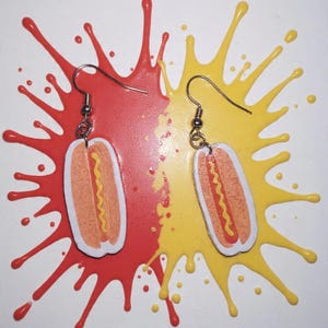 Könnte beinhalten: Ein Paar ausgefallene Ohrringe mit Hotdog-Design. Jeder Ohrring hat einen weißen Umriss, ein hellorangefarbenes Brötchen, einen roten Hotdog und einen gelben Senfstreifen. Die Ohrringe hängen an silberfarbenen Haken, vor einem roten und gelben Farbspritzer.