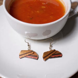 Könnte beinhalten: Ohrringe im Design eines gegrillten Käsesandwiches. Die Ohrringe sind hellbraun mit dunkelbraunen Grillstreifen und einer roten Füllung, die an silberfarbenen Haken hängen. Die Ohrringe werden auf einem weißen Teller präsentiert.