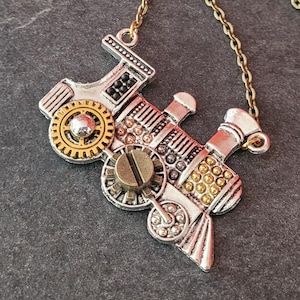 May include: A steampunk-style pendant necklace featuring a detailed train engine design. The pendant is silver-toned with gold and bronze accents, including gears, rivets, and a screw head. The necklace has a bronze-colored chain.
