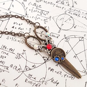 Puede incluir: Un collar de bronce con un colgante de tijeras antiguas. Las tijeras tienen un adorno de piedras preciosas rojas y azules y una esfera de reloj con números romanos. El collar está sobre un fondo blanco con ecuaciones matemáticas y diagramas.