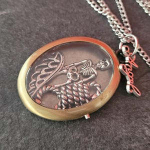 Puede incluir: Collar con colgante ovalado con un diseño de esqueleto plateado dentro de un recinto de vidrio transparente, enmarcado por metal dorado. El colgante cuelga de una cadena plateada, con un dije de "Angel".