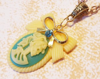May include: A gold chain necklace with a blue and white cameo pendant featuring a woman's profile. The pendant is accented with a gold bow and a small blue rhinestone.