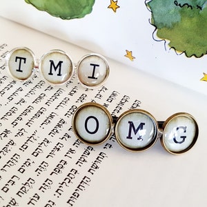 May include: A set of six typewriter key charms spelling out "TMI OMG". The charms are silver and gold with white backgrounds and black lettering.
