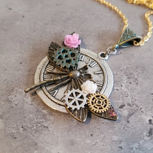 May include: A silver necklace with a dragonfly pendant featuring a clock face, gears, and pink and white flowers.