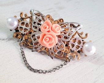 May include: A decorative barrette with a steampunk design. It features a silver filigree butterfly, gears, and peach-coloured roses. Two faux pearls and a chain add detail to this unique hair accessory.