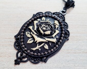 May include: Black and cream-colored rose pendant necklace. The pendant features a detailed rose design set against a black background, framed by a decorative black border with small bead accents. The necklace has a black chain.