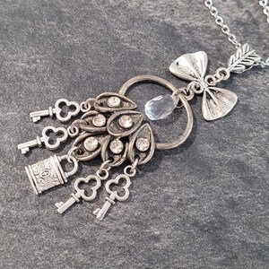 Op de afbeelding: Zilverkleurige hangerketting met een cluster van sleutels en een slotbedel. De hanger heeft een centraal ontwerp met heldere kristallen accenten en een druppelvormige kristal. Een pijl-en-boogbedel staat bovenaan.