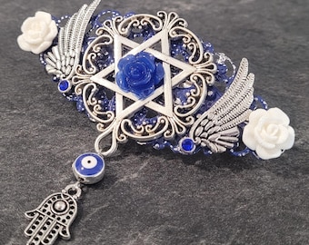 May include: A silver-toned hair barrette with a blue and white color scheme. The barrette features a Star of David, a blue rose, white roses, and silver wings. A dangling charm includes an evil eye and a Hamsa hand.