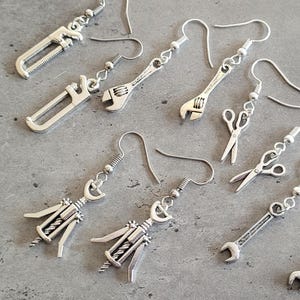 May include: Silver-toned earrings featuring miniature tools, including a saw, adjustable wrench, scissors, and corkscrew. The earrings have a detailed design and are suspended from simple ear wires. The collection is displayed on a gray surface.