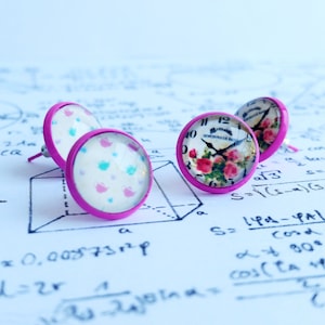 May include: A set of three pink stud earrings with different designs. One earring features a floral pattern with a clock face, another has a pattern of birds, and the third has a floral pattern.
