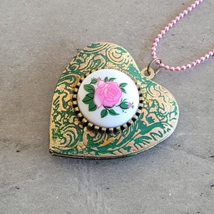 Könnte beinhalten: Eine herzförmige Halskette mit einem Anhänger, der ein rosafarbenes Rosenmotiv auf weißem Hintergrund zeigt. Der Anhänger ist grün und goldfarben mit einem detaillierten Muster. Die Kette ist rosa.