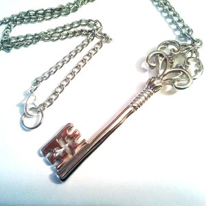 May include: Silver key pendant necklace. The key features an ornate top and a straight stem. The necklace chain is silver and has a clasp. The key is approximately 7.6 cm long.