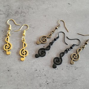 Puede incluir: Tres pares de pendientes con un diseño de clave de sol. Un par es dorado, uno es negro y uno es bronce.