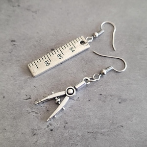 May include: Silver-toned earrings, one shaped like a ruler with the numbers 84, 85, and 86, and the other a compass. The earrings have a hook closure. The earrings are on a gray surface.