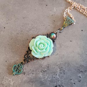 May include: A mint green rose charm pendant necklace with an antique bronze filigree design and a key charm. The necklace is on a silver chain.