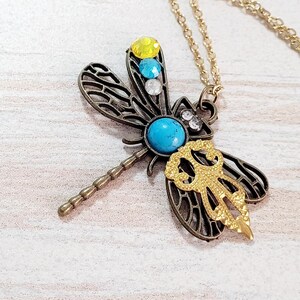 May include: A dragonfly pendant necklace with a gold-tone chain. The dragonfly has bronze-colored filigree wings, a turquoise center stone, and yellow, blue, and clear accent stones. A gold-colored decorative element is on the dragonfly's body.