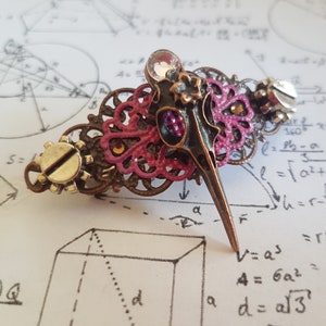 Könnte beinhalten: Eine Haarklammer aus rosa und bronzefarbenem Metall mit einem Vogelmotiv. Der Vogel hat einen langen Schnabel und ein detailliertes Design. Die Klammer ist mit kleinen, klaren Strasssteinen verziert.
