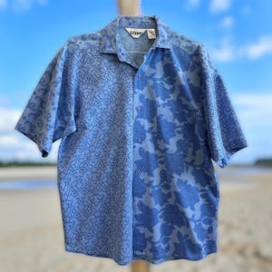 May include: A light blue button-down shirt with short sleeves. The shirt features a split design, with a paisley pattern on the left side and a floral pattern on the right side. The shirt is hanging on a hanger.