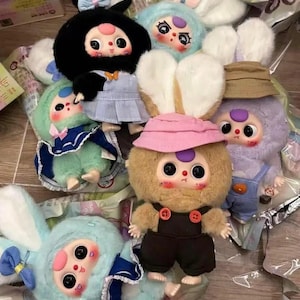 May include: A collection of plush bunny dolls with large eyes and rosy cheeks. The dolls are in various colors, including black, blue, green, and purple, and wear outfits like overalls and dresses. Some have hats and bows.