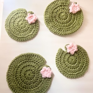 May include: Four handmade crochet coasters. Each coaster is a circular shape in a light green color, resembling a lily pad. A small pink flower is attached to each coaster. The coasters are designed to protect surfaces from drink condensation.