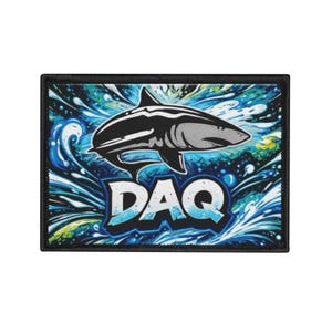Peut inclure: Écusson rectangulaire avec une bordure noire, représentant un requin stylisé en noir et gris sur un fond d'éclaboussures d'eau bleues et blanches. Le mot "DAQ" est affiché en blanc et bleu. Design audacieux.
