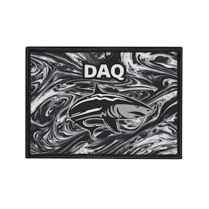 Peut inclure: Un écusson rectangulaire avec une bordure noire. L'écusson présente un fond tourbillonnant noir et blanc avec une illustration de requin au centre. Le mot "DAQ" est imprimé en blanc en haut de l'écusson.