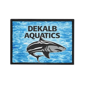 Peut inclure: Un écusson rectangulaire avec un motif de requin. Le requin est noir et blanc, sur un fond d'eau bleue. Le texte "DEKALB AQUATICS" est affiché en lettres blanches au-dessus du requin.