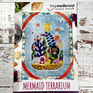 Peut inclure: Un modèle de point de croix compté représentant une sirène dans une bouteille en verre avec du corail, des algues et d'autres créatures marines. Le motif est intitulé "Mermaid Terrarium".