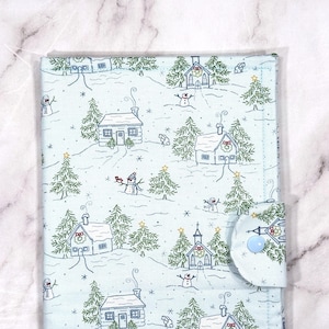 May include: A light blue fabric item featuring a winter village design. The pattern includes houses, snowmen, and fir trees. A light blue button secures the flap. The item is likely a case or cover.