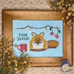 May include: Cross stitch pattern featuring a fox with a mug of hot cocoa and the text "Cozy Season". The pattern is framed in a wooden frame and set against a blue background.