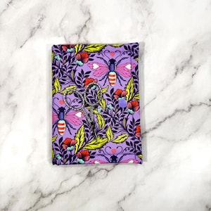 May include: A purple fabric scissor case with a floral and bee pattern. The case holds a pair of small, silver scissors. The design includes pink bees, red flowers, and yellow leaves. A purple button secures the case.