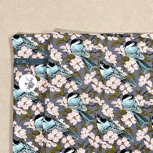 May include: A grey fabric featuring a repeating pattern of blue and black birds and pink flowers. A zip is visible on the left side, with a round tag attached. The tag has a paw print design. The fabric is likely used for a bag or pouch.