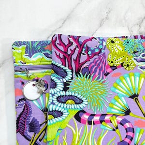 May include: A fabric pouch featuring a vibrant underwater scene with purple, lime green, and teal coral, sea anemones, and a seahorse. The pouch includes a purple zip closure with a silver-toned circular charm attached to the zip pull.