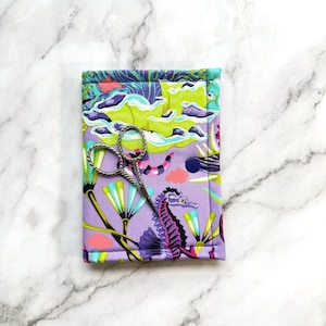 May include: A rectangular fabric case featuring a vibrant pattern of purple seahorses, green seaweed, and coral accents, secured with a dark blue snap button. A pair of silver metal embroidery scissors with a twisted handle rests on the case.